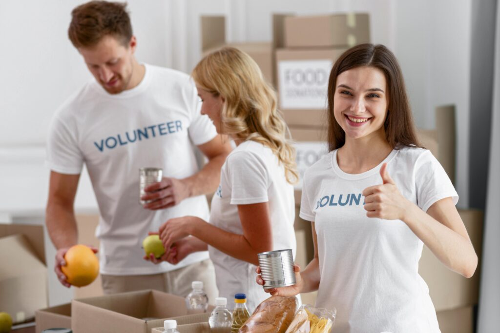 Three volunteers pack food items into boxes. A woman in the foreground holds a can and gives a thumbs-up.